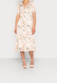 Robe midi florale dans un pastel clair avec manches courtes, col en V, détails à boutons, et coupe ample, complétée par des sandales blanches à brides.