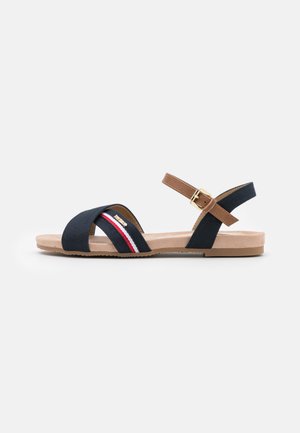 Flat navy blue sandal with crossed front straps, brown ankle strap with gold buckle, red and white stripe detail on side strap.