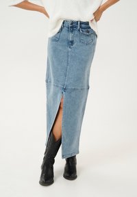 Jeans-Maxikleid mit einem vorderen Schlitz, hellblaue Farbe, klassisches Fünf-Taschen-Design und kontrastierende Nähte. Getragen mit schwarzen Cowboystiefeln.