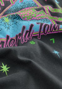 Schwarzer Stoff mit lebhaftem blauen, rosa und grünen Grafikdruck, der Leopardenmuster, Sterne und den Text "Weltreise" in einer fetten Schriftart zeigt.