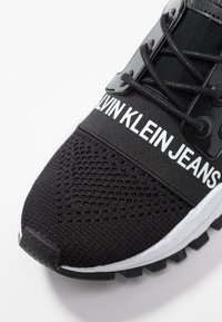 Svart stickad sneaker med robust sula, svarta snörning och vit "Calvin Klein Jeans" band över toppen.