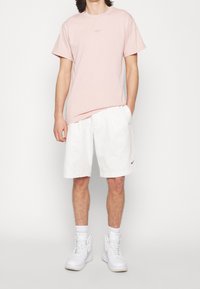 Světle růžové bavlněné tričko s malým logem na hrudi, doplněné bílými bavlněnými šortkami. Oblečení doplňují bílé sneakers s vysokou podrážkou.