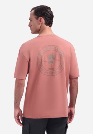 Man met kort, donker haar die een zalmroze T-shirt draagt met een cirkelvormige palmboomgrafiek en de tekst "Mysterious Impermanence" op de rug.