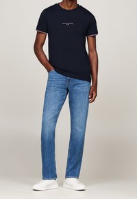 Navy cotton t-shirt with red and white trim, featuring a small logo on the chest. Paired with light blue denim jeans and white sneakers.