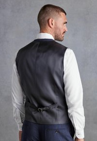 Chaleco de satén negro con una textura suave, que presenta una hebilla trasera única y una camisa blanca a juego, colocado sobre un fondo gris liso.