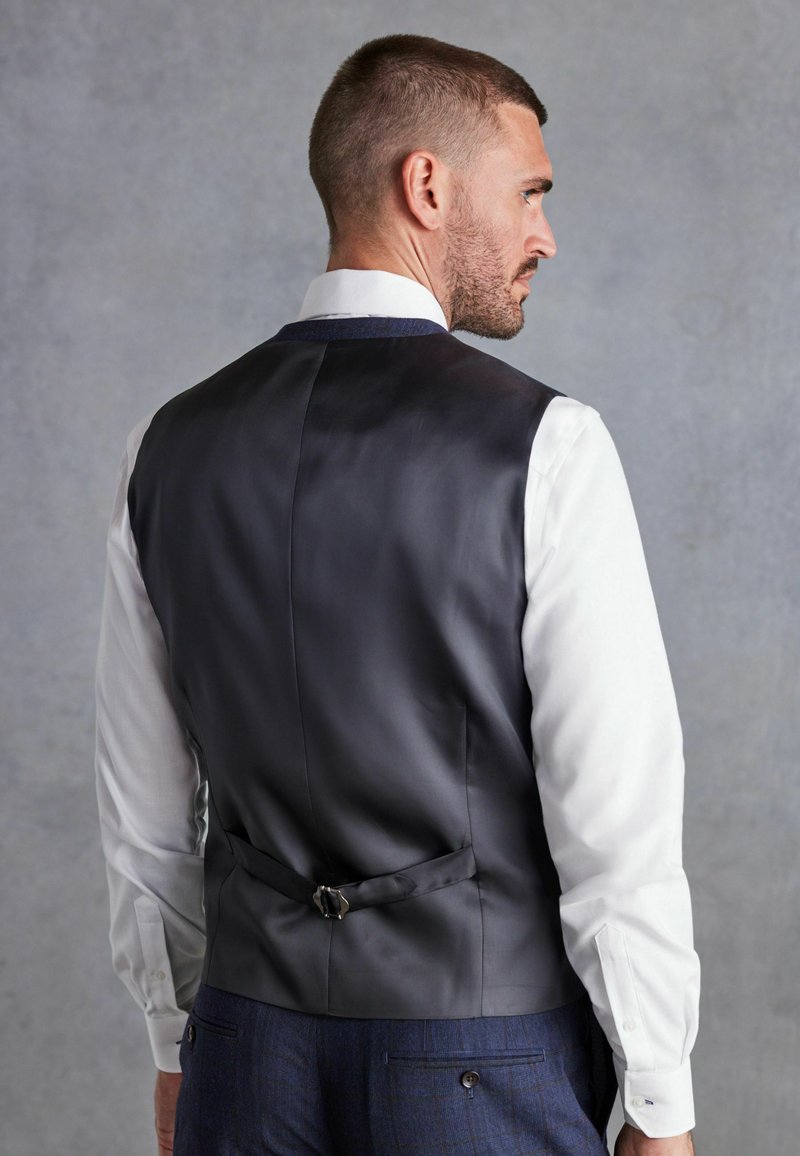 Chaleco de satén negro con una textura suave, que presenta una hebilla trasera única y una camisa blanca a juego, colocado sobre un fondo gris liso.