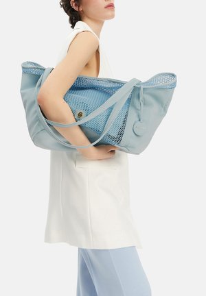Kobieta trzyma na ramieniu dużą jasno-niebieską siatkową torbę typu tote, ubrana w bezrękawnik biały i jasno-niebieskie spodnie.
