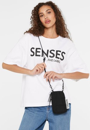 Weißes oversized T-Shirt mit schwarzem Text, kombiniert mit blauen Jeans. Eine kleine schwarze Umhängetasche aus strukturiertem Material mit verstellbarem Tragegurt.