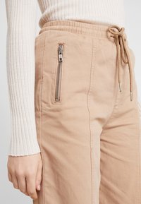 Beżowe spodnie cargo z elastycznym pasem, sznurkiem i boczną kieszenią na zamek. Miękka tkanina o gładkiej strukturze i zwężanym kroju.
