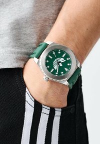Montre avec un bracelet en silicone vert, lunette argentée, cadran vert et index heures blancs, portée au poignet, partiellement visible contre un pantalon noir.