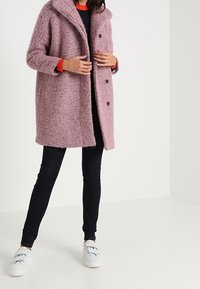 Femme ajustant un manteau rose texturé boutonné, portant un pantalon noir et des baskets blanches, debout devant un fond clair uni.