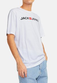 Weißes Baumwoll-T-Shirt mit schwarzem Text "JACK & JONES" und rotem rechteckigen Akzent. Lockere Passform und kurze Ärmel, kombiniert mit hellblauen Jeans.