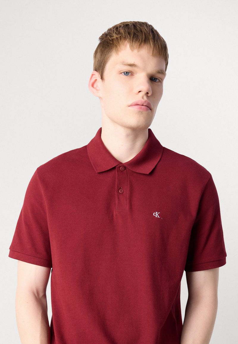 Bordowa koszulka polo z bawełny, z kołnierzykiem, plakatką z trzema guzikami i małym logo po lewej stronie klatki piersiowej. Krótkie rękawy.