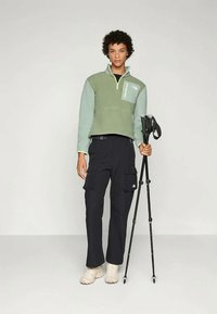 Fleece-pullover i myk grønn med glidelås hals, med en brystlomme; parret med mørke cargo-bukser og beige turbots. Går med stav.