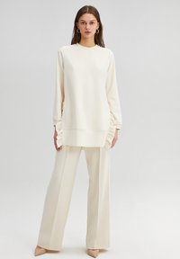 Long-sleeve off-white top with textured ruffle details at the sides, paired with loose off-white trousers, complemented by beige pointed shoes.