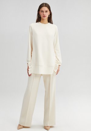 Dlhý rukáv off-white top s textúrovanými volánovými detailmi po stranách, kombinovaný s voľnými off-white nohavicami, doplnený béžovými špicatými topánkami.