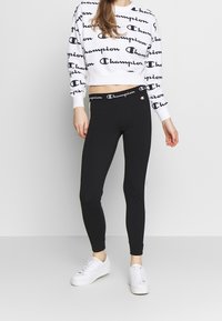 Kobieta w czarnych legginsach z paskiem w logo Champion, białej bluzie Champion z czarnymi logo oraz białymi trampkami, stojąca na szarej podłodze.