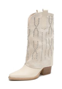 Bota de cowboy beige con parte superior de ante texturizado, adornada con tachuelas plateadas en varios patrones, y un tacón bajo apilado para mayor estabilidad.
