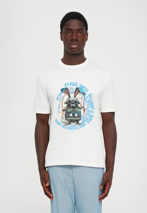 Homme portant un t-shirt blanc avec un lapin robot vintage coloré tenant une cassette, associé à un pantalon bleu clair à rayures.