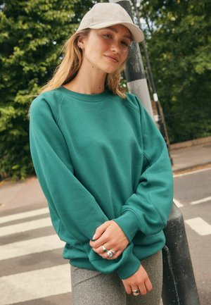 Grüner Sweatshirt mit lässigem Schnitt, gerippten Bündchen und rundem Ausschnitt. Kombination mit strukturierten grauen Hosen und einer beigen Baseballcap.