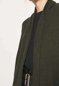 Femme portant un cardigan texturé vert olive sur un haut noir, assorti d'une ceinture noire avec une boucle rectangulaire dorée.