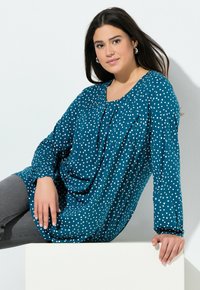 Vrouw met lang donker haar draagt een blauw patroon blouse en grijze broek, casual zittend op een wit blok tegen een effen achtergrond.
