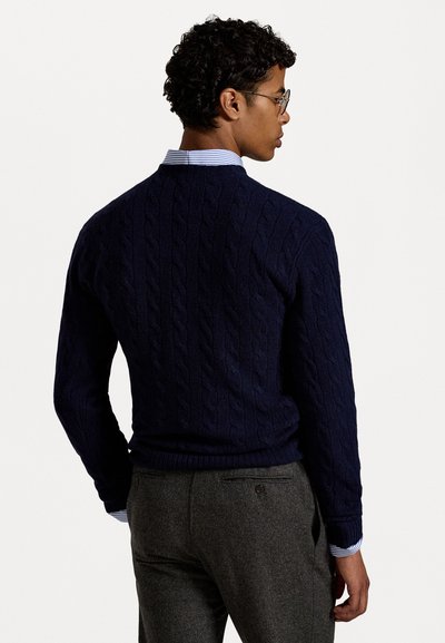 Navy-Strickpullover mit Rippbündchen und Saum, getragen über einem hellblauen gestreiften Hemd, kombiniert mit dunkelgrauen Hosen.