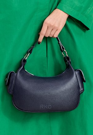 Dłoń z czarnym lakierem do paznokci trzyma małą czarną teksturowaną skórzaną torebkę z metalowymi pierścieniami na uchwycie na tle zielonego płaszcza.