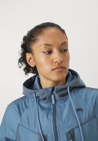 Casaco azul funcional com capuz, frente com fecho éclair e texturas contrastantes, com detalhe de logotipo e cordões ajustáveis.