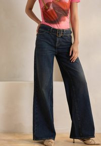 Breda ben mörk denimjeans med hög midja, försedda med ett stort bälte och subtila blekningar. Kombineras med en rosa grafisk t-shirt.