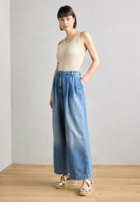 Top beige senza maniche, pantaloni in denim azzurro chiaro a vita alta e larga, e sandali espadrilles dorati. Caratteristiche includono una vita con cintura e pieghe.