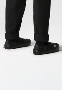 Black padded slip-on shoes with a textured surface and logo detail, paired with rolled black pants and black socks.