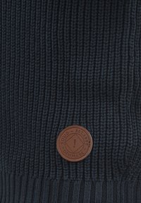 Navy gestrickte Mütze mit gerippter Textur und einem runden braunen Lederpatch, das den Markennamen und ein Logo zeigt.