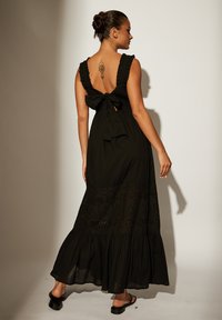 Svart maxiklänning med ett rynkat halslinne och knytning i ryggen. Har en djupare skuren rygg och mönstrad fåll. Slät tygkonstruktion med en flödande siluett.