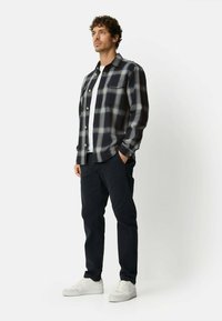 Koszula w czarno-szarą kratę z kieszeniami na piersi, pod spodem biała koszulka, ciemne spodnie i białe trampki. Postawa stojąca na jednolitym tle.