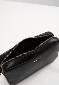 Czarna torba z teksturowanej skóry z złotym zamkiem błyskawicznym, posiadająca jedną przegrodę i wytłoczone inicjały marki na przedniej stronie.