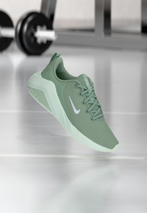 Chaussure de course Nike verte avec logo swoosh blanc, flottant dans une salle de sport avec des poids flous en arrière-plan.