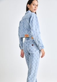 Chaqueta y pantalones de mezclilla celeste con un patrón de logo DKNY en toda la prenda, con etiqueta distintiva y pequeño acento bordado en el bolsillo trasero.
