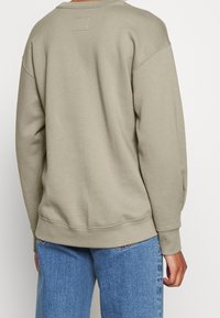 Sudadera verde oliva hecha de tela suave, con cuello redondo, hombros caídos y puños acanalados con una etiqueta rectangular en la parte trasera.