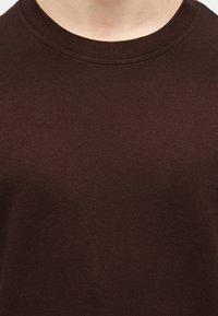 Κοντινό πλάνο ενός ατόμου που φοράει μια απλή σκουρόχρωμη καφέ βαμβακερή μπλούζα με στρογγυλή λαιμόκοψη, εμφανίζοντας το λαιμό και το πάνω μέρος του στήθους.