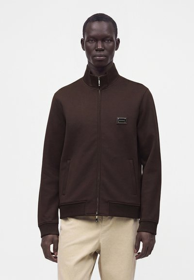 Homme portant une veste zippée brun foncé avec un petit patch sur la poitrine et un pantalon beige clair, debout devant un fond blanc uni.