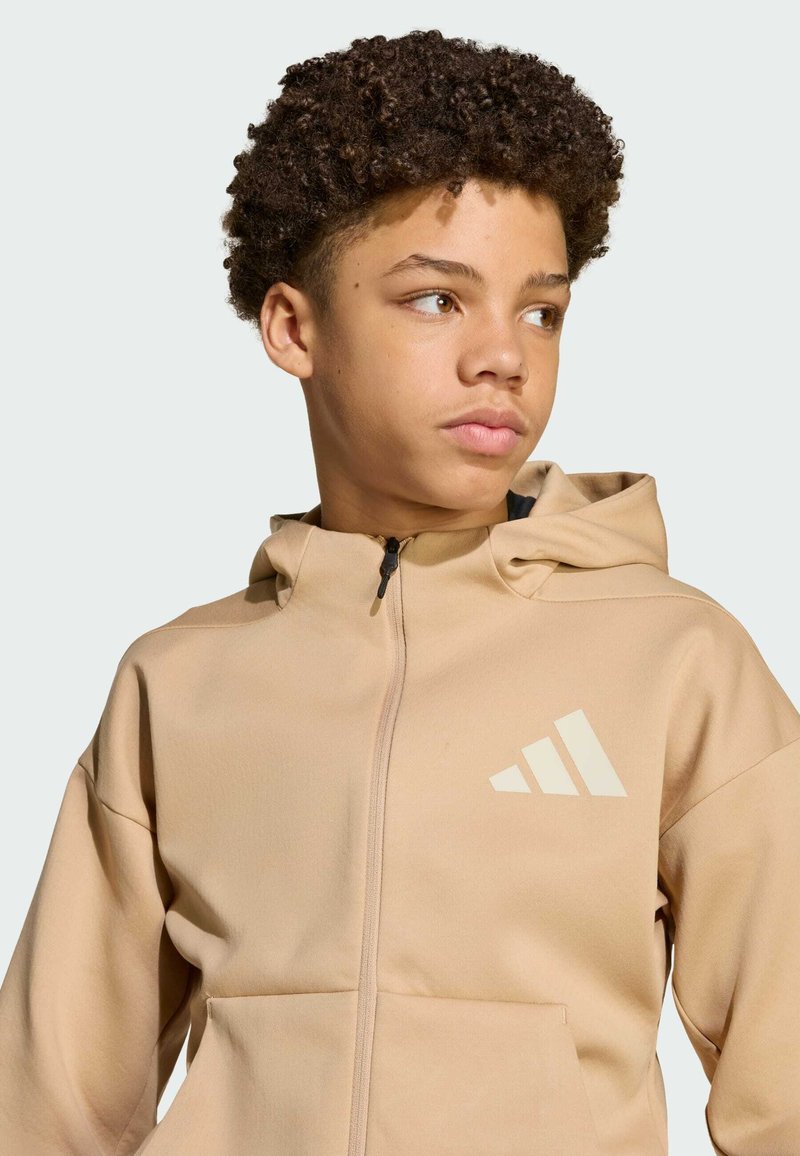 Pješčana zip majica s kapuljačom, koja se odlikuje glatkom teksturom i logotipom u svjetlijoj bež boji. Uključuje prednje džepove i prilagođeni kroj.