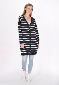 Navyblauwe en witte gestreepte cardigan met een V-hals, knoopsluiting aan de voorkant en lange mouwen, gecombineerd met lichtblauwe spijkerbroek en witte sneakers.
