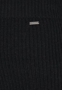 Schwarzer Strickstoff mit gerippter Textur, versehen mit einem kleinen silbernen Metallschild, das "DreiMaster" eingeprägt hat. Einfaches und einheitliches Design.