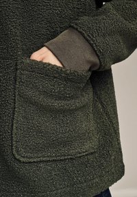 Fuzzy green fleece jacket with a large front pocket and ribbed cuff. The texture is soft, plush, and richly detailed.