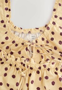 Gelber Stoff mit kastanienbraunen Polka-Dots, der elastisch geraffte Kanten und Bindebänder zeigt, dargestellt sind der Ausschnitt- und der Taillenbereich eines Kleidungsstücks.