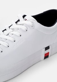 Weißes Leder-Sneaker mit glatter Oberfläche, ausgestattet mit einer Gummisohle, flachen weißen Schnürsenkeln und einem gestreiften Akzent in Rot, Weiß und Blau an der Ferse.