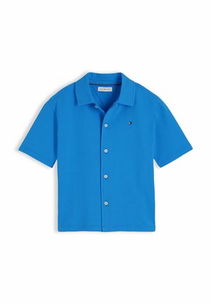 Chłopiec stoi i się uśmiecha, nosi niebieską koszulę z krótkim rękawem i guziczkami, czarne szorty, białe skarpetki i czarne mokasyny na białym tle.