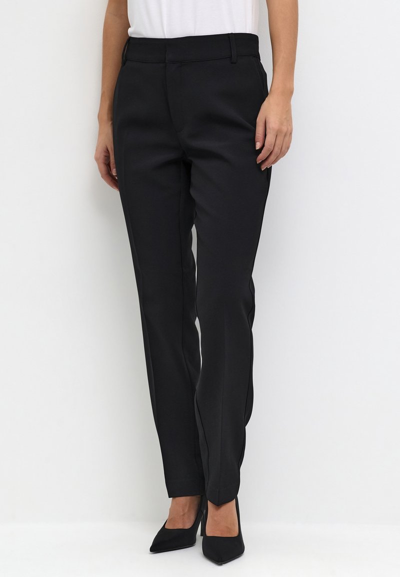 Femme portant un pantalon noir ajusté avec un pli à l'avant, associé à des talons noirs pointus, debout sur un fond blanc.