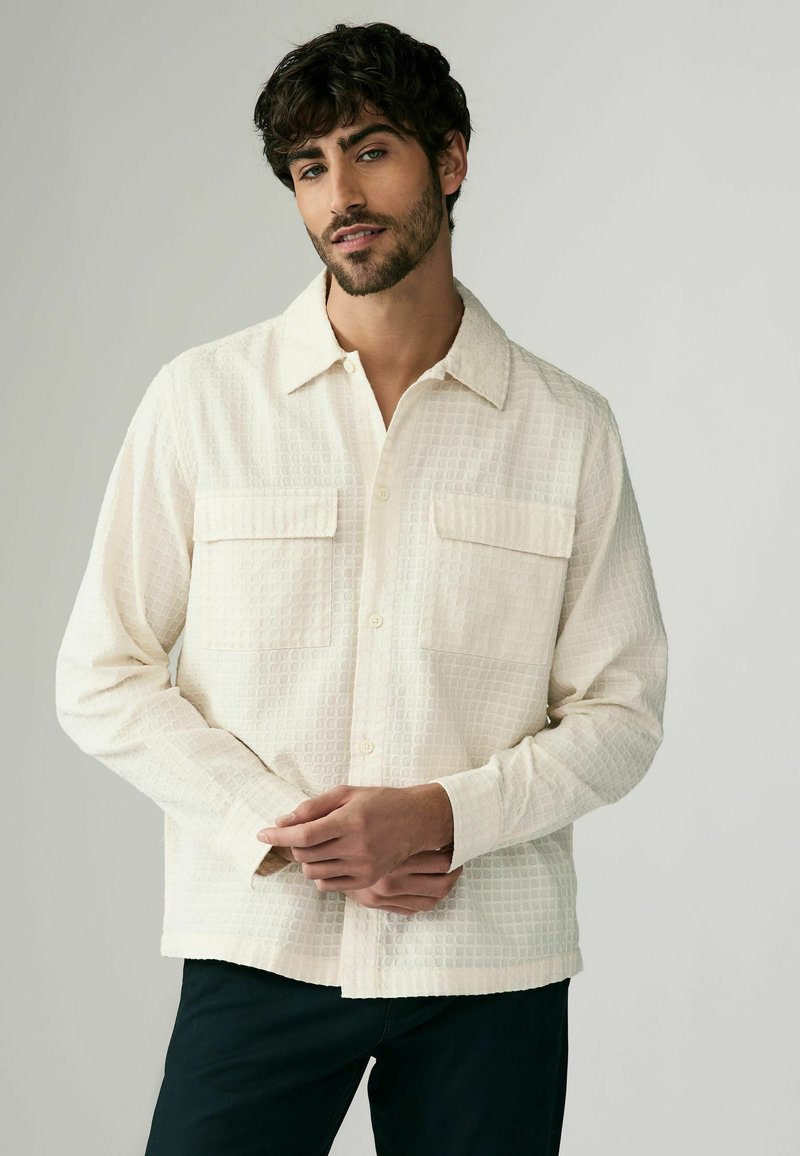 Beige button-up shirt with a textured grid pattern, featuring two chest pockets and a classic collar. Slim fit with long sleeves.